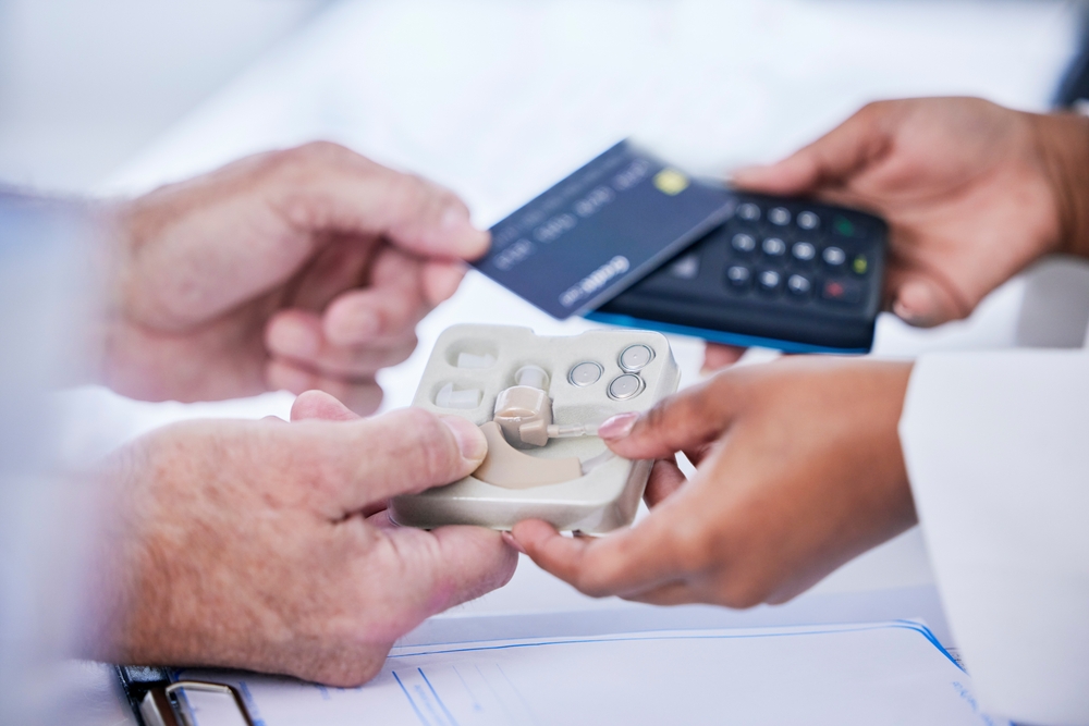 A customer makes a payment with a credit card using a portable card reader while receiving a hearing aid device from a healthcare professional.