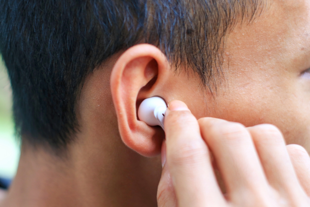 A close-up of a person wearing a white wireless earbud in their ear, with one hand adjusting the earbud. The side of the persons head and ear are clearly visible.
