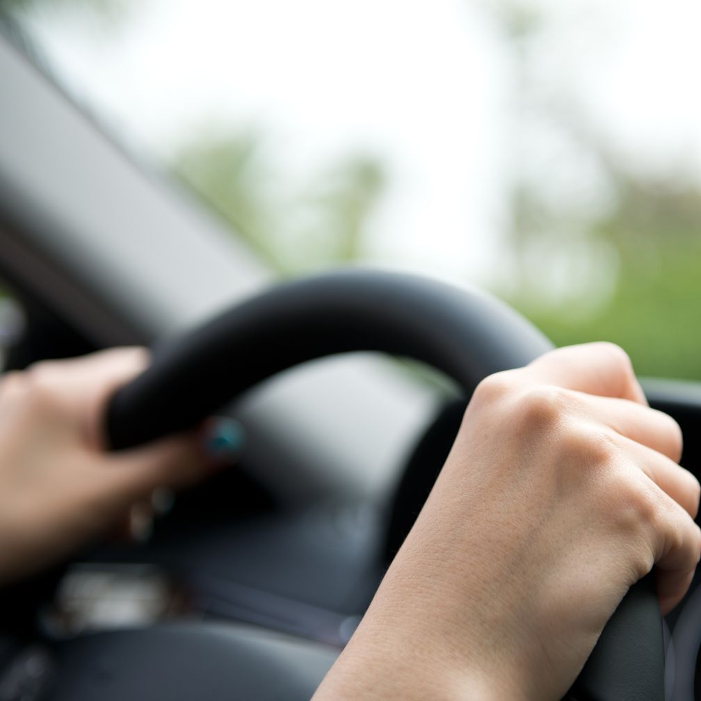 Close-up of two hands gripping a black steering wheel, with a blurred background visible through the car window, suggesting the person is driving.