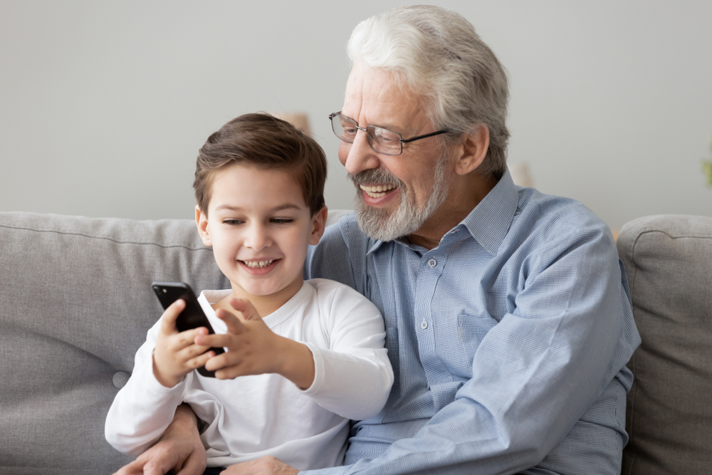Smiling elderly man with gray hair and beard sits on a couch with a young boy. The boy is holding a smartphone and both are looking at it, appearing happy and engaged.