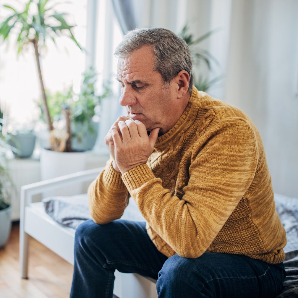An older man in a mustard sweater sits on a bed, leaning forward with hands clasped, looking thoughtful or concerned. Indoor plants and soft natural light are visible in the background.