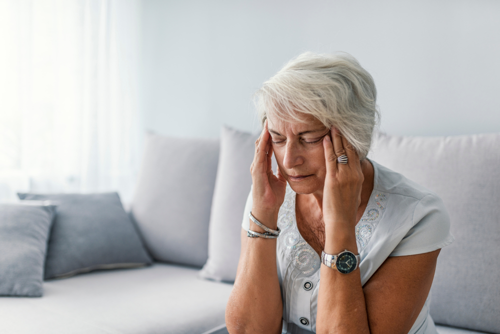 An older woman with short gray hair sits on a sofa, eyes closed, and holds her temples with both hands, appearing to be in pain or discomfort.