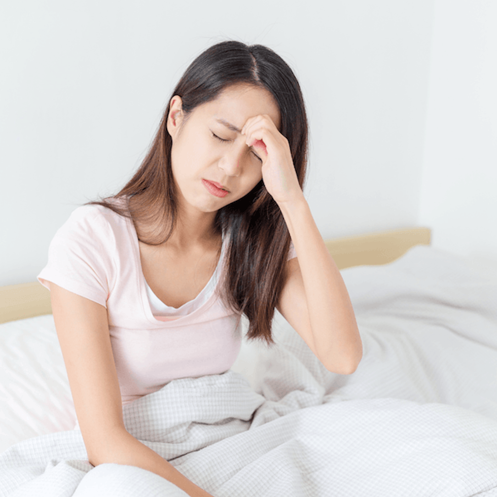 A woman sits in bed with her hand on her forehead, eyes closed, appearing to have a headache or feel unwell.