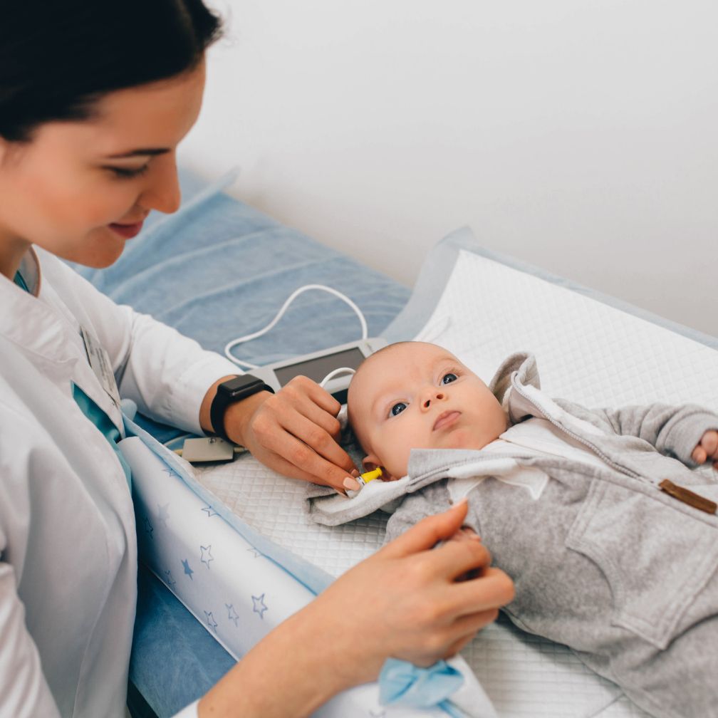 A healthcare professional tests a babys hearing using medical equipment while the baby lies on an examination table covered with a white mat.