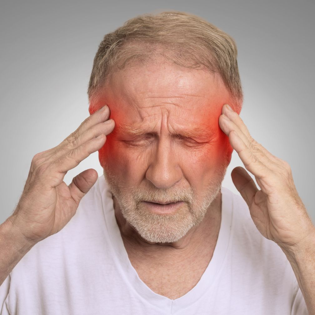 Older man with closed eyes holds his temples, showing discomfort. Red highlights on both sides of his head indicate pain, suggesting he may have a headache or migraine. He wears a white shirt and stands against a gray background.