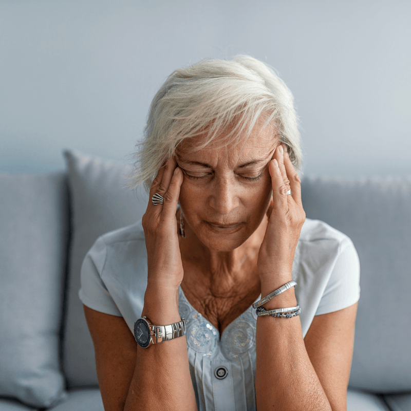 Older woman with short white hair sits on a couch, eyes closed, pressing her temples with both hands, appearing to have a headache or stress. She wears a light blue top and silver jewelry.