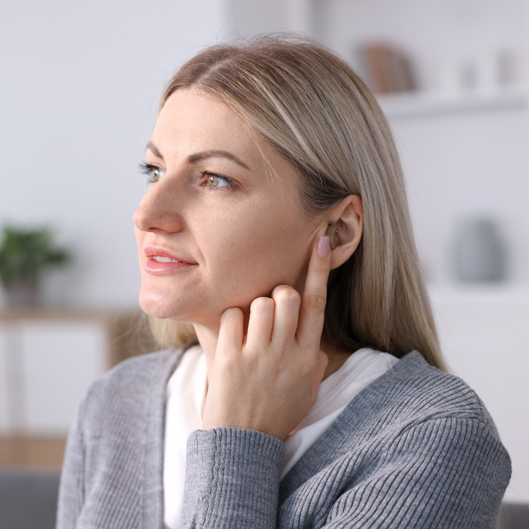 A woman with blonde hair touches her ear with her finger while looking to the side, wearing a gray sweater, indoors with a blurred background.