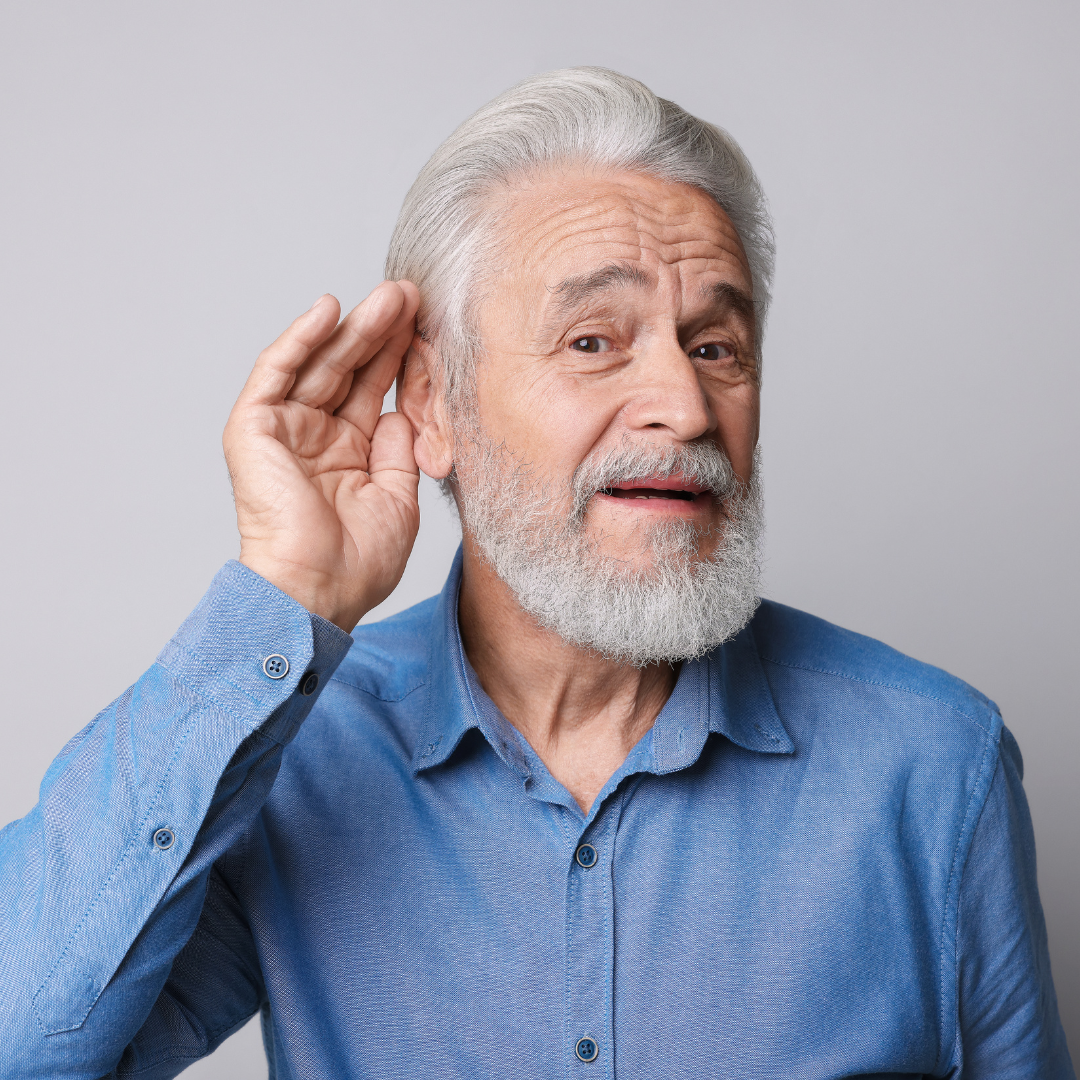 Older man with gray hair and beard wearing a blue shirt, holding his hand to his ear and tilting his head as if trying to hear better, standing against a plain light gray background.